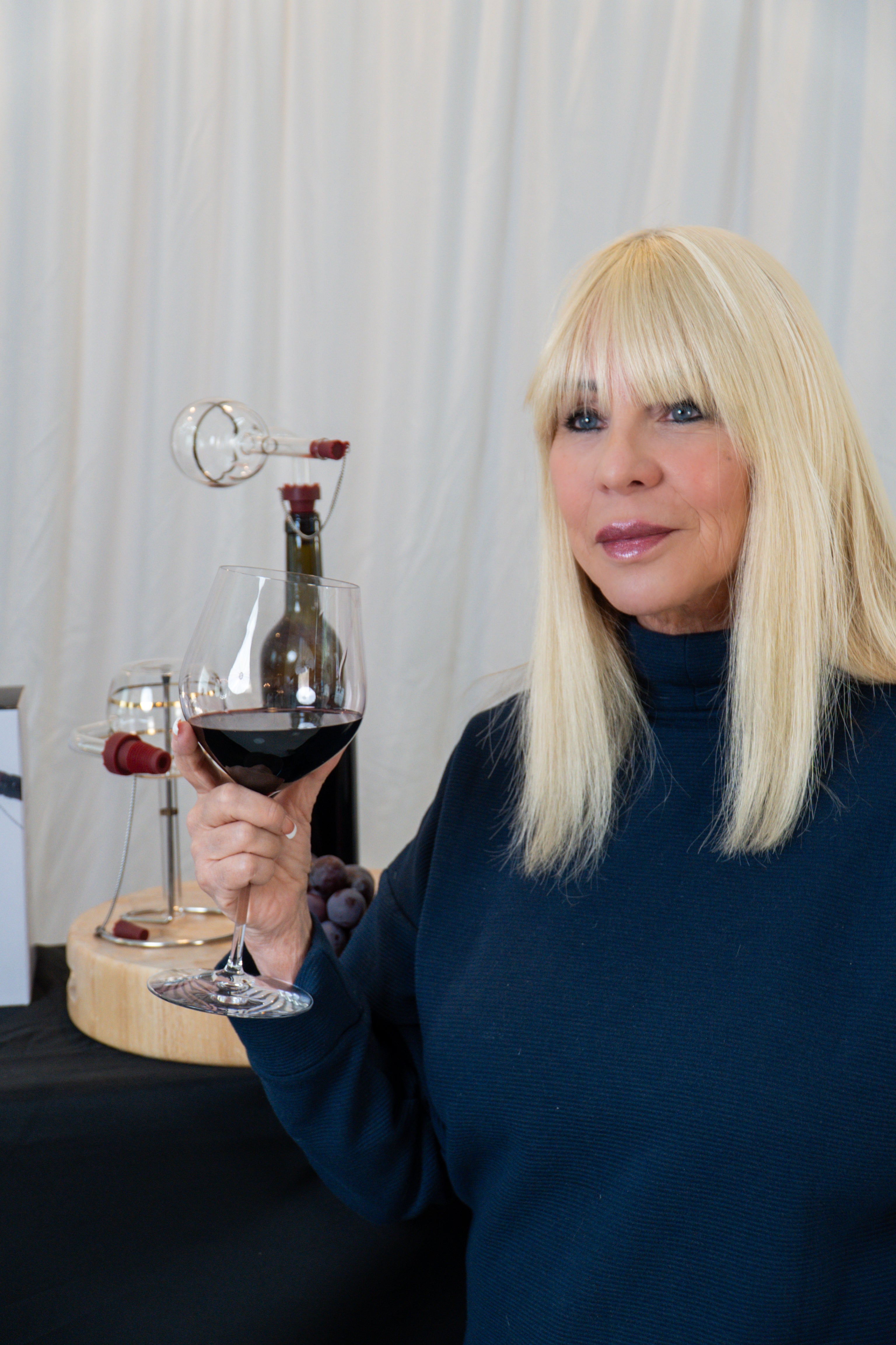 A blonde woman in a navy blue turtleneck sweater holds a glass of red wine in front of a white curtain backdrop, with a wine bottle and glassware on a table beside her.